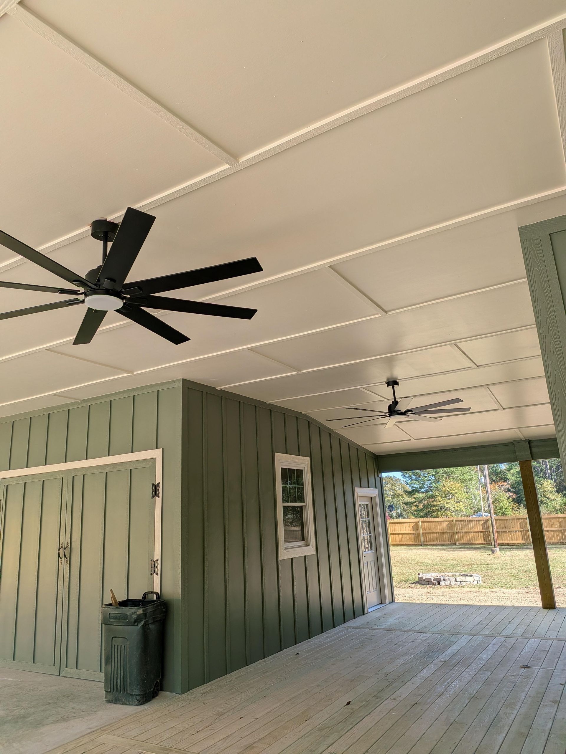 Covered porch with green siding, white ceiling, black ceiling fans, and a trash bin near the wall.
