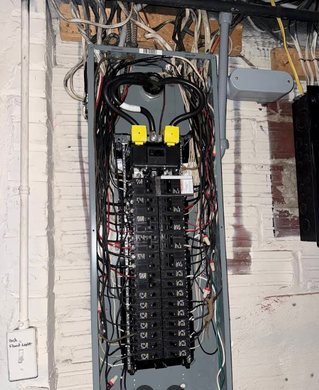 Electrical breaker panel with tangled wires in a utility room, mounted on a wall.