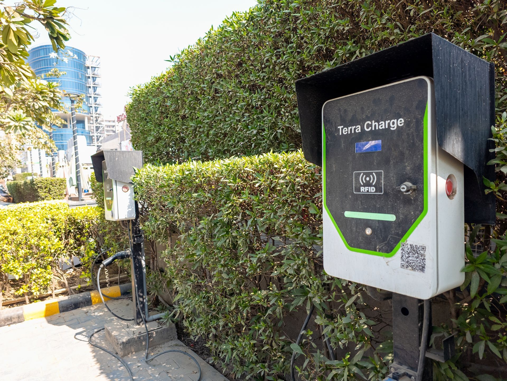 Outdoor EV charging station beside a hedge, with a black-and-white charger cabinet and cable on a sunny sidewalk.