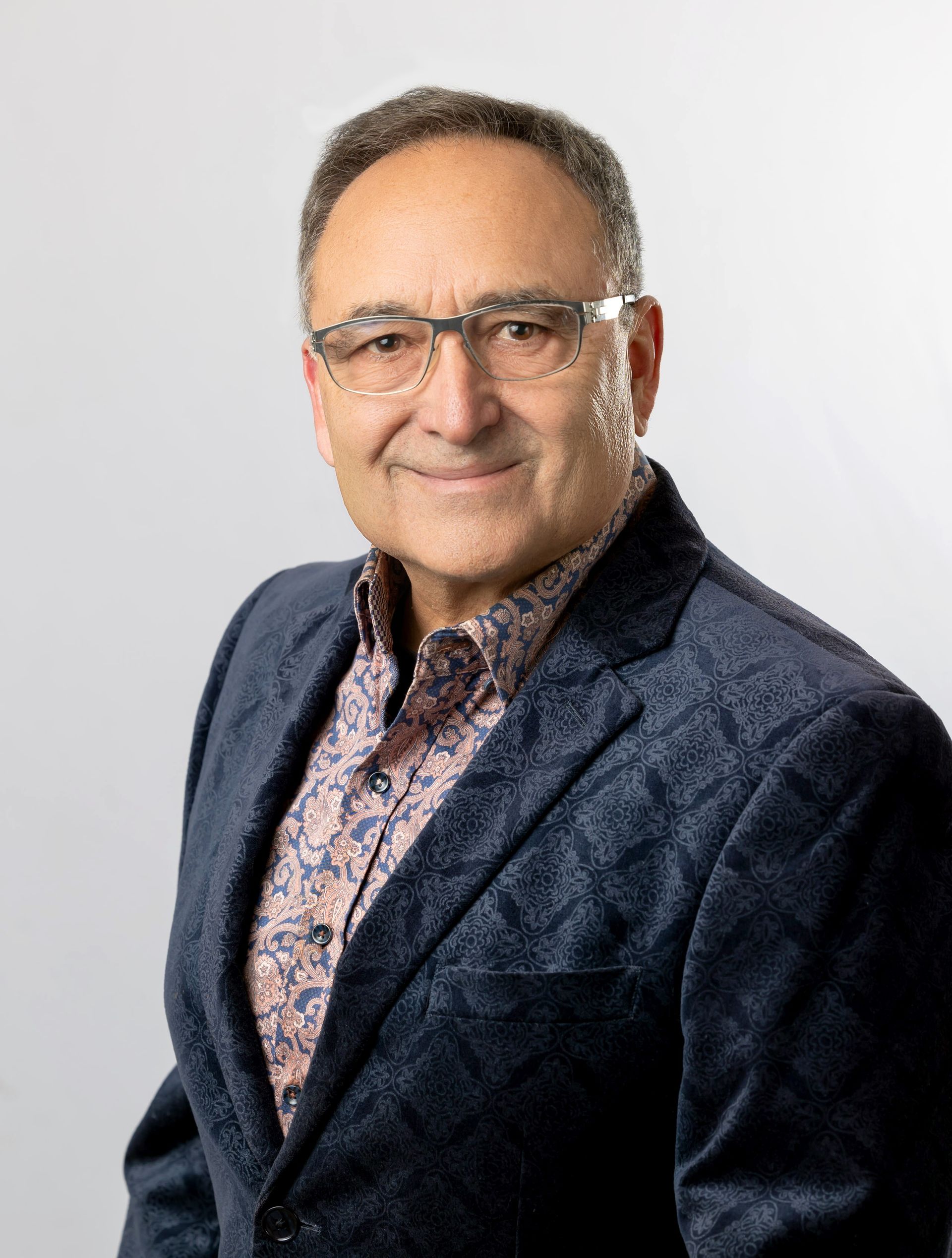 Man with glasses wearing a patterned shirt and blue jacket, smiling, studio shot.