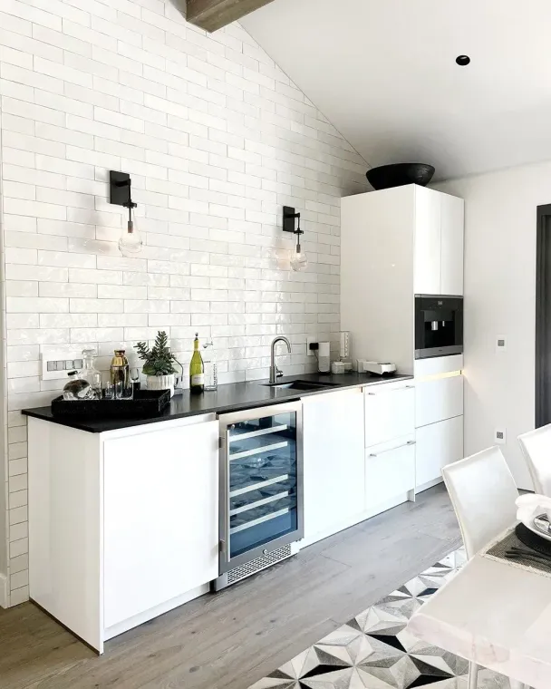 White modern kitchen with wine fridge, black countertops, brick wall, and a light-colored floor.
