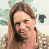 אישה עם שיער חום בהיר ארוך וחיוך קל, לובשת חולצה עם דוגמאות על רקע צורות ירוקות ושחורות.