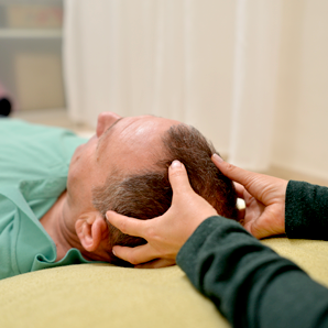 אדם המקבל עיסוי ראש שוכב על מזרן צהוב. ידיו מונחות על ראשו של האדם.