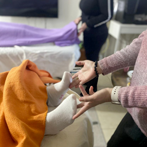 רגלי אדם מכוסות בשמיכה כתומה וגרביים לבנות בזמן ששני אנשים מבצעים טיפול ריפוי; ידיים מוחזקות מעל כפות הרגליים.