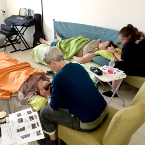 אנשים מקבלים עיסוי ראש בחדר. שני אנשים שוכבים על מיטות, בעוד שניים אחרים מעסים את ראשיהם. ברקע נמצא כיסא מתקפל.