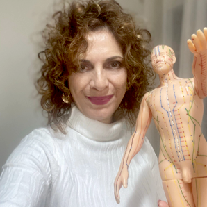 אישה עם שיער חום מתולתל וצווארון גולף לבן, אוחזת בדמות אנטומית גברית המציגה מרידיאנים של דיקור.