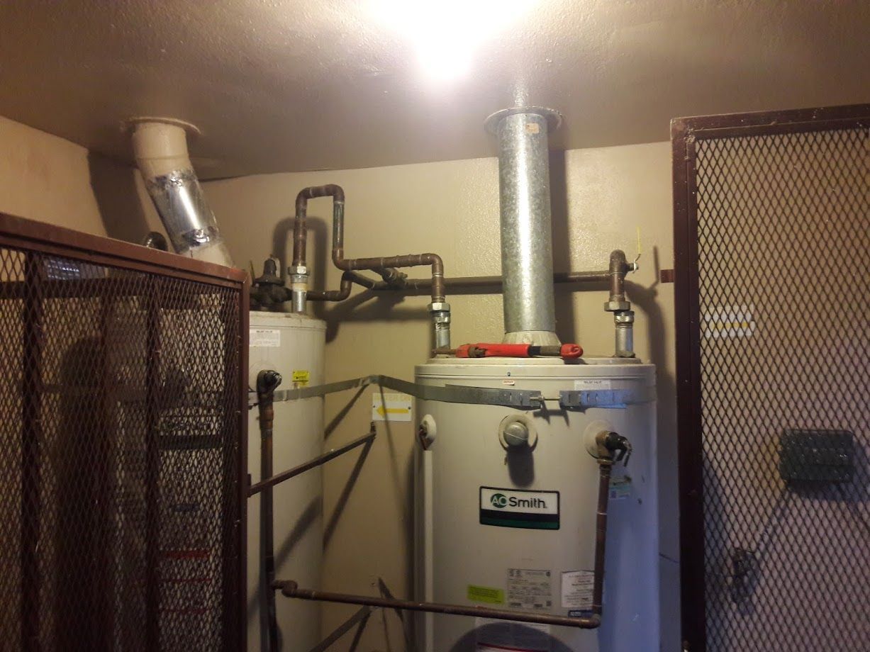 Two water heaters in a utility room, with metal venting and piping. Metal grate surrounds the space.