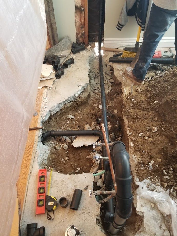 Plumbing work in progress: Trench cut in concrete floor, black pipes and copper fittings. A person stands nearby.