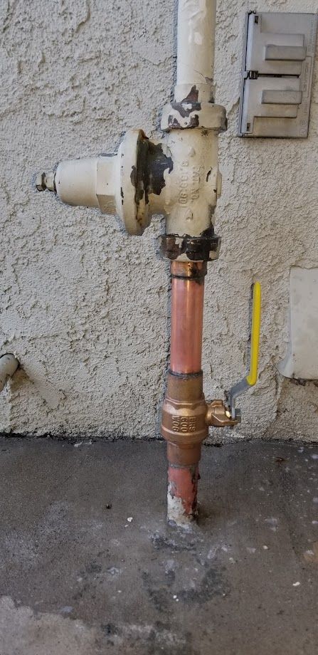 A rusty water pressure regulator with copper and white pipes attached to the wall.