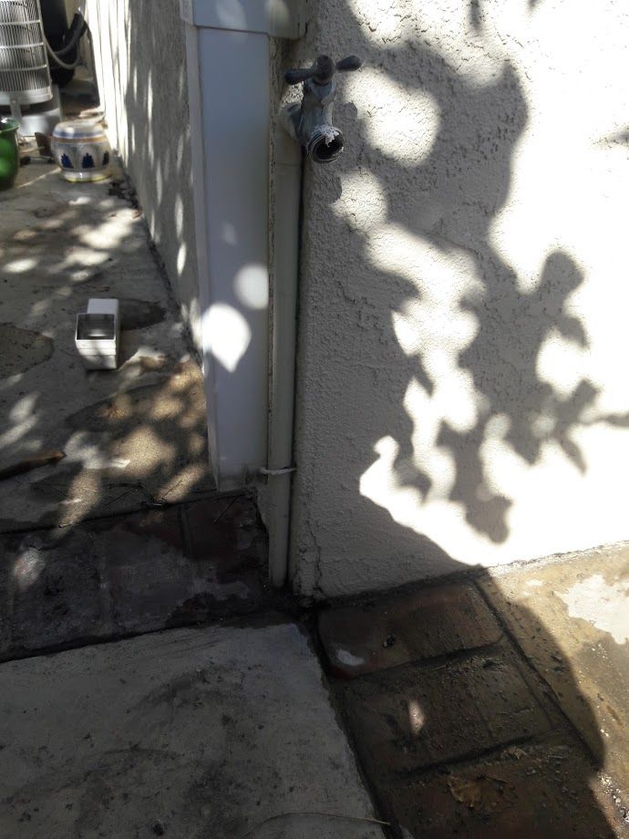 White pipe and faucet on a stucco wall, with shadows cast across the concrete ground.