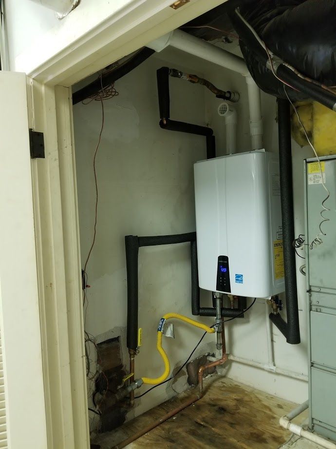 A small utility closet with a white tankless water heater, black pipes, and yellow gas line.