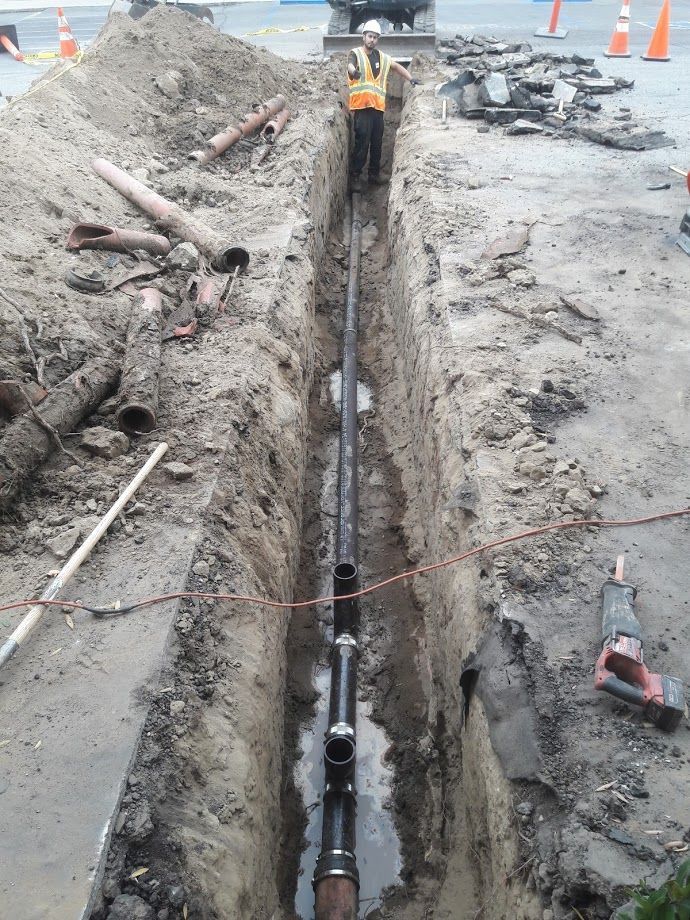Construction worker in trench, installing black pipe. Tools and debris surround.