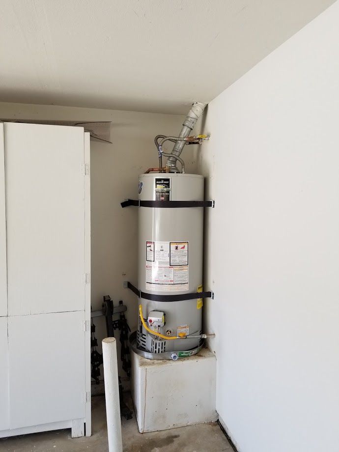 A water heater in a garage, secured with straps. It has vents, pipes, and is near white walls and a cabinet.