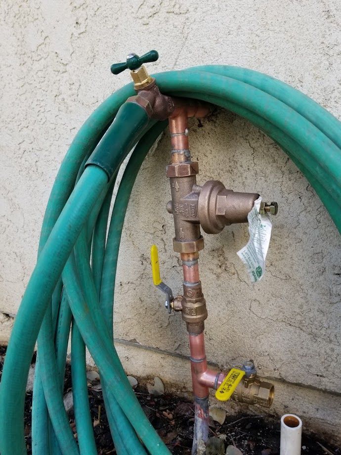 Copper plumbing with hose and valves attached to a wall. Green hose, brass fittings, and yellow-handled valves.