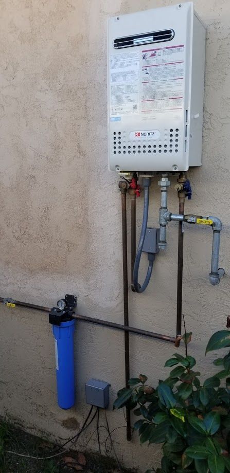 A wall-mounted, white tankless water heater with plumbing, a blue water filter, and greenery.