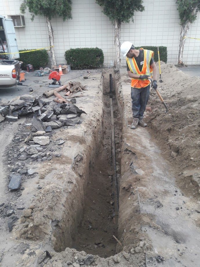 Construction worker in hard hat inspecting trench; asphalt debris, building in background.