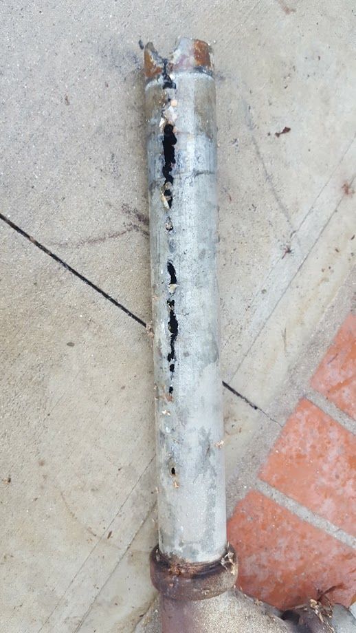 Corroded metal pipe with black streaks and broken top, resting on a concrete surface.