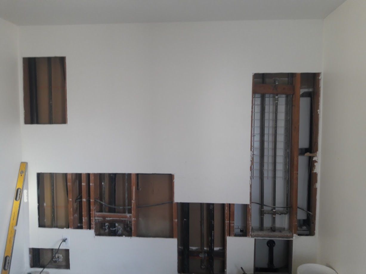 White wall with multiple rectangular sections cut out, exposing wood framing and pipes.