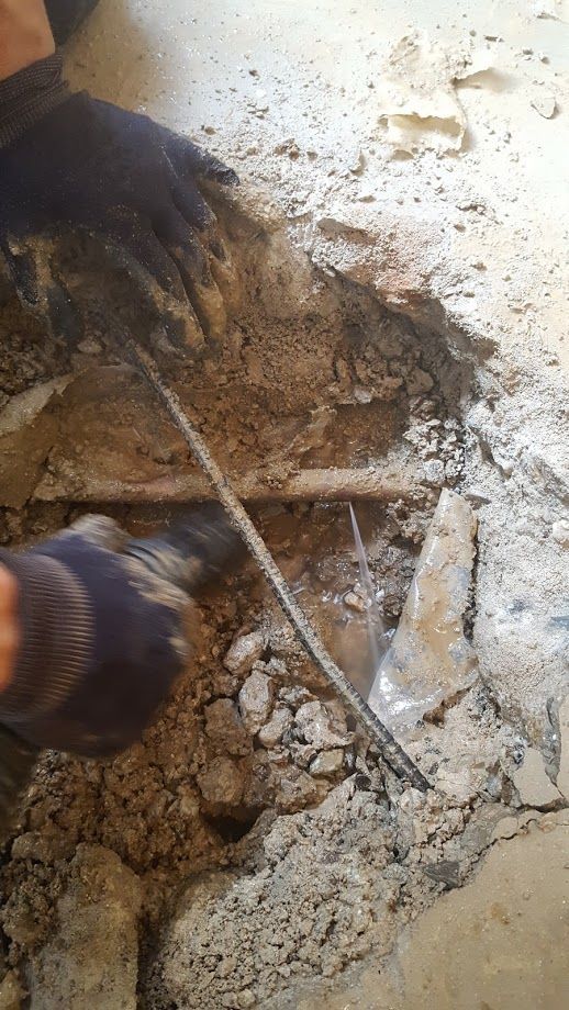 Hands wearing gloves excavating concrete, revealing metal pipes in a construction setting.