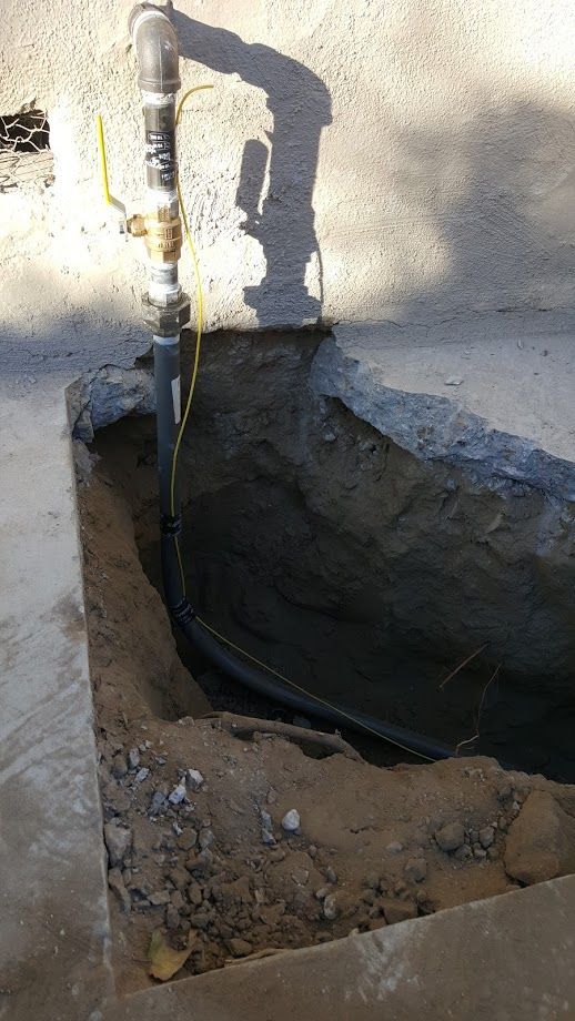 Trench dug next to a concrete surface, revealing a black pipe connected to a vertical gas line.