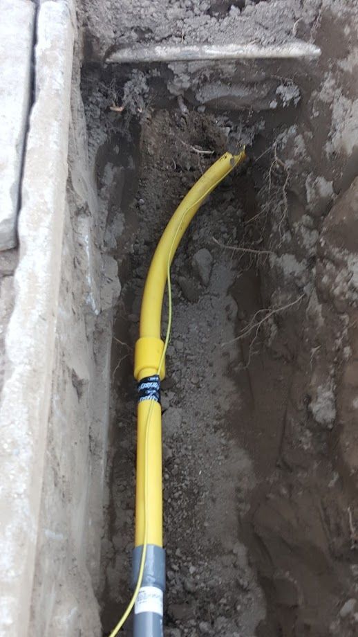 Yellow gas line in a trench, connected to a pipe. Soil and concrete borders the line.