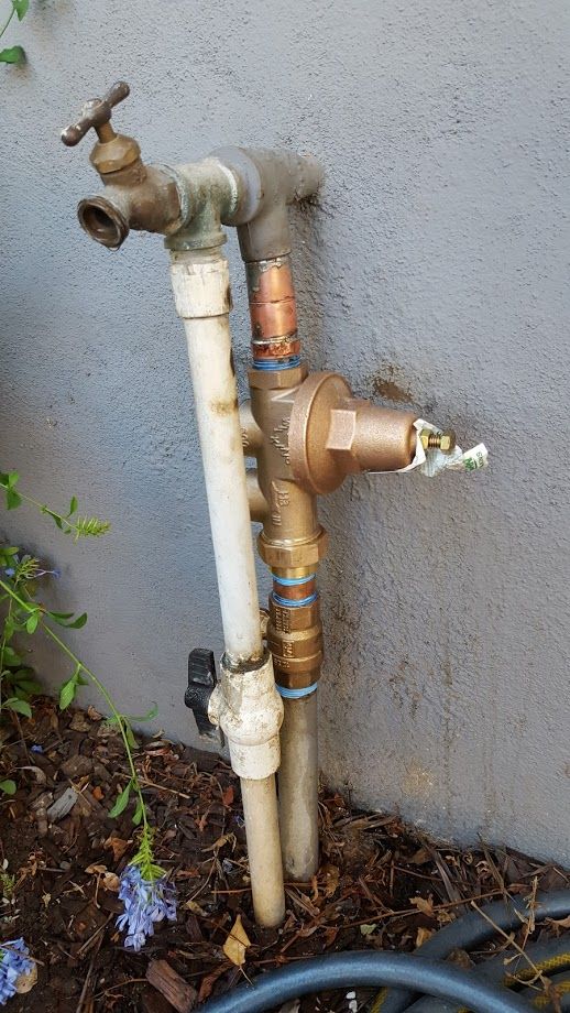 Outdoor water pipes and fixtures against a stucco wall, including a faucet, pressure regulator, and PVC pipes.