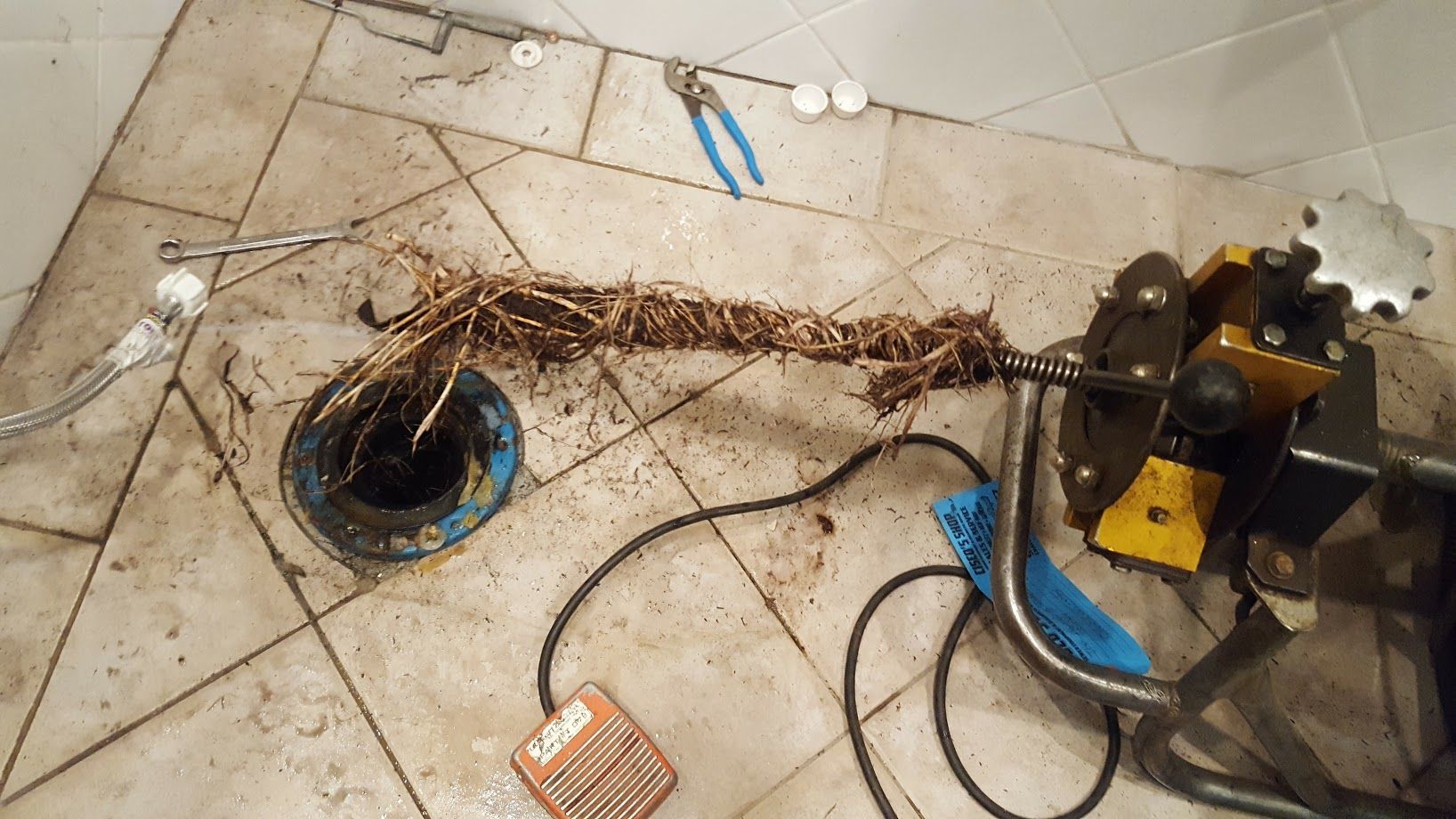 A plumbing snake removes roots from a drain. Messy bathroom floor, tools visible.
