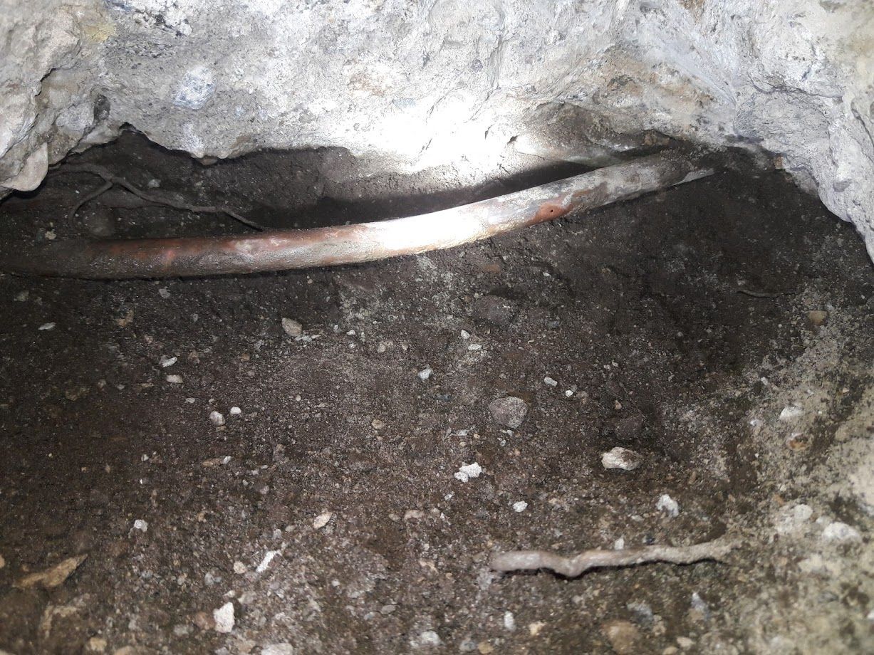 Copper pipe exposed in a hole in concrete. Dark, wet soil surrounds the pipe.