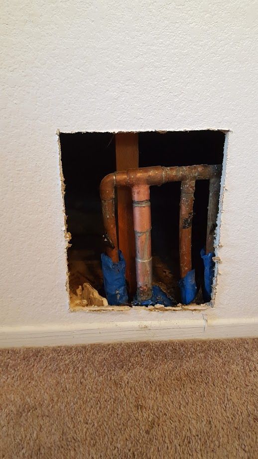 Hole in a white wall exposing copper pipes with blue insulation, close to a beige carpet.