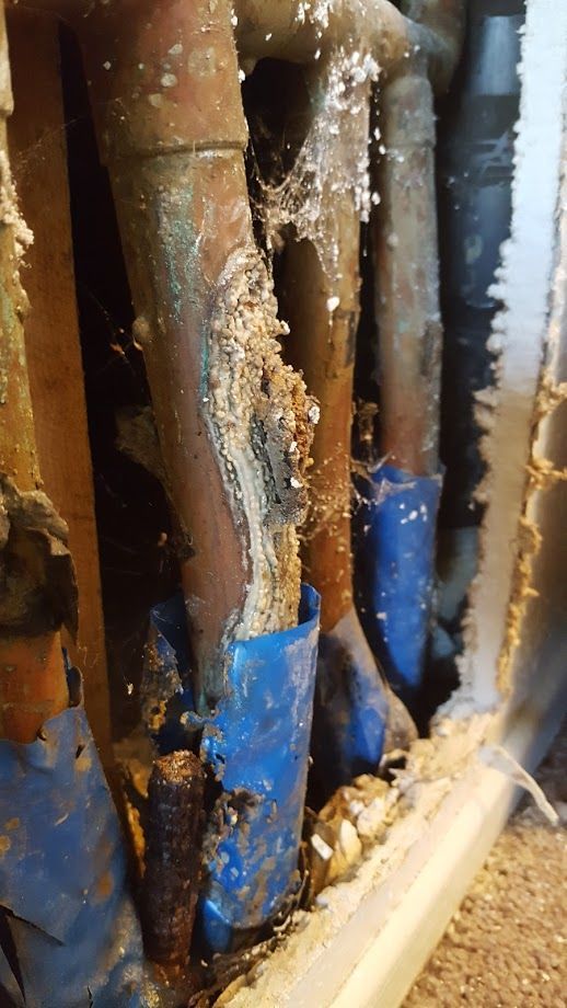 Corroded copper pipes with white mineral buildup and blue tape in a wall cavity.