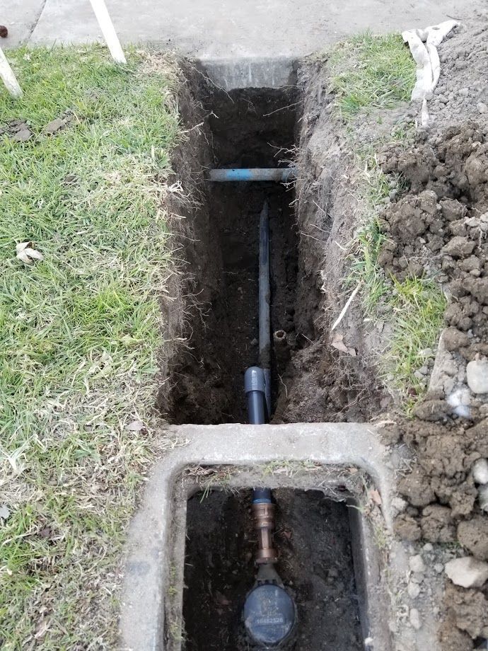 Trench dug in grass, exposing water meter and pipes. Dark soil, gray concrete, and a blue pipe are visible.