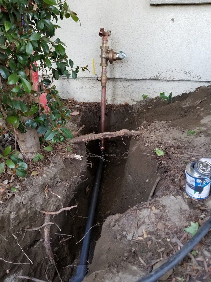 Dug trench near a building. Copper pipe and water valve are visible. Black pipe runs through the trench.