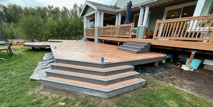 A large, multi-level wooden deck attached to a house, featuring gray-bordered stairs leading down to a grassy backyard.