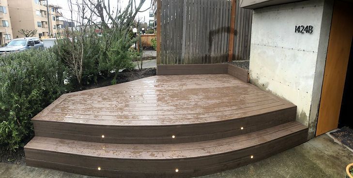 A two-tiered wooden deck with built-in lights, situated outside a building with a privacy screen and surrounding greenery.