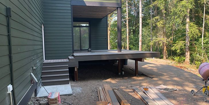 A dark-sided home with a raised wooden deck and stairs under construction, set in a wooded area with gravel ground.