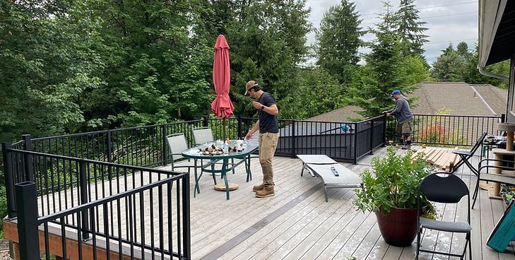 A person stands on a wooden deck with black railings, a table, a red umbrella, and chairs overlooking trees and houses.