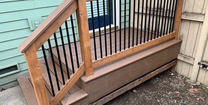 A wooden porch with dark railings and a small set of stairs leading to a doorway, set against a light green wall.