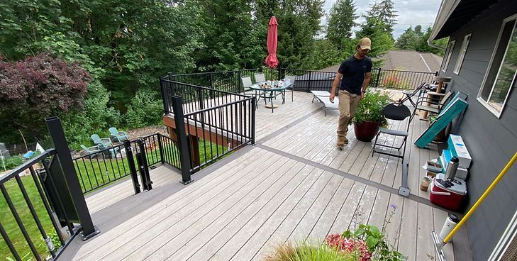 A person walks across a light gray wooden deck with black railings, overlooking a grassy yard with trees.