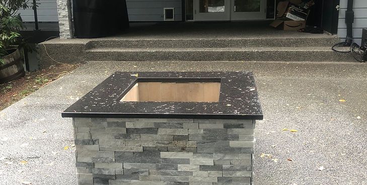 A square, stone-tiled fire pit with a black countertop, situated on a concrete patio in front of house steps.