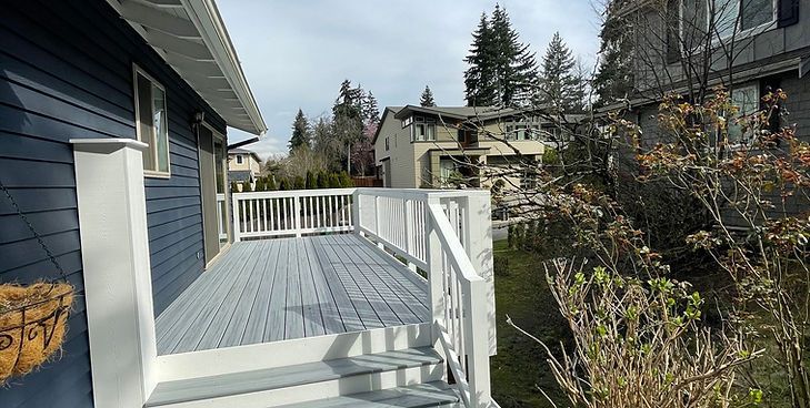 A newly painted light gray deck with white railings attached to a blue house, viewed from the steps on a sunny day.