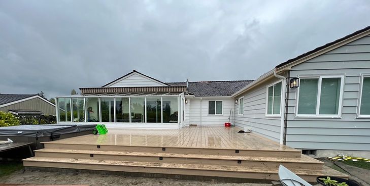 A light gray house features a newly built wooden deck with wide steps leading to a glass-enclosed sunroom under a cloudy sky.