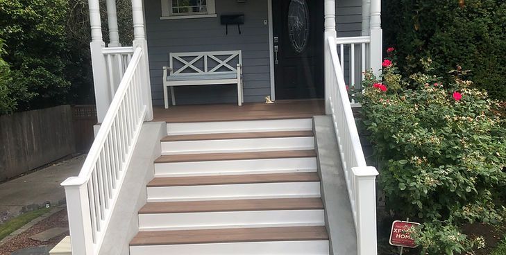 Stairs with white railings leading up to a porch with a gray wall, a white bench, and a red rose bush to the right.