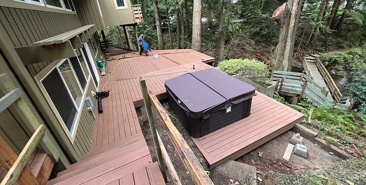 A person works on a new wood-colored deck containing a covered hot tub, situated near a house in a wooded area.