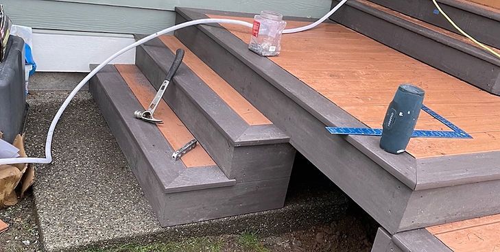 A hammer, a square, and a container of fasteners sit on unfinished wood-toned steps with grey trim during construction.