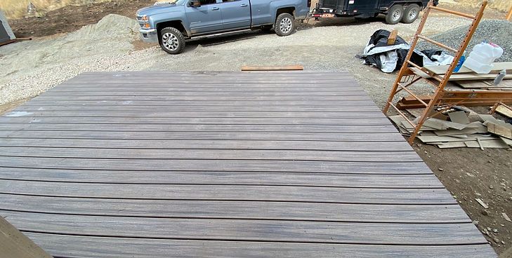 A rectangular deck made of weathered grey wooden planks, set in an outdoor construction area with a truck and trailer nearby.