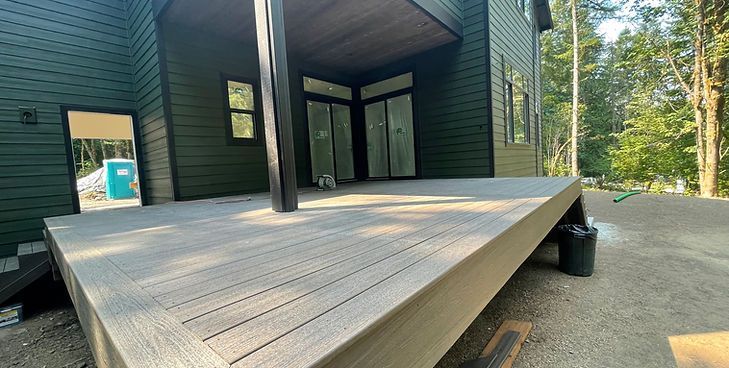 A large, light-colored wooden deck attached to a modern house with dark green horizontal siding and sliding glass doors.