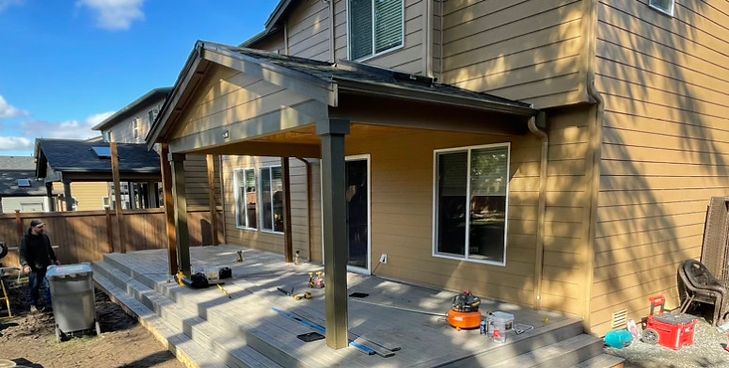 A tan two-story house with a newly constructed covered patio featuring a dark wooden frame and a concrete tiered deck.