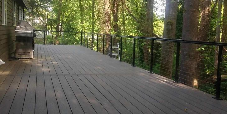 A wooden deck with a black railing, overlooking a wooded area with a grill on the side.