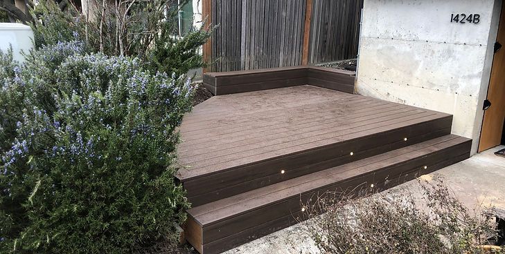 A two-step dark brown wooden deck sits in front of a white wall next to a large, leafy shrub.