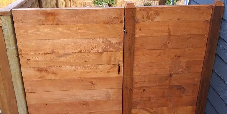 A double wooden gate with horizontal planks, stained in a warm brown, set between a fence post and a house wall.